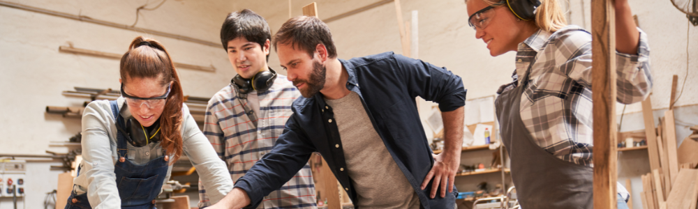 Banner image of an employer showing three apprentices how to use a tool in a carpentry workshop.